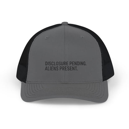 Gray and black trucker hat with 'DISCLOSURE PENDING. ALIENS PRESENT.' text and cross symbol on a white background
