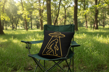 A square black throw pillow featuring a gold line-art graphic of a Bigfoot silhouette walking beneath a flying saucer, shown on a clean white background.