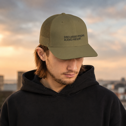 A man wearing a dark olive green trucker cap with black embroidered text reading 'DISCLOSURE PENDING. ALIENS PRESENT.